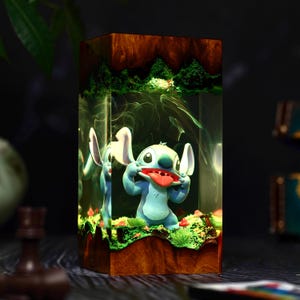 May include: A rectangular resin art piece featuring a blue cartoon character with large ears and a red tongue. The character is set within a clear resin block, surrounded by a miniature landscape with green foliage and a wooden base.