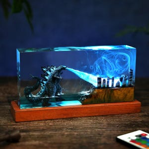 Könnte beinhalten: Harzskulptur mit Godzilla, der eine Stadt mit einem blauen Strahl angreift. Die detaillierte Figur ist in einem transparenten Block eingeschlossen und auf einem Holzsockel montiert. Die Szene wird mit blauem Licht beleuchtet.