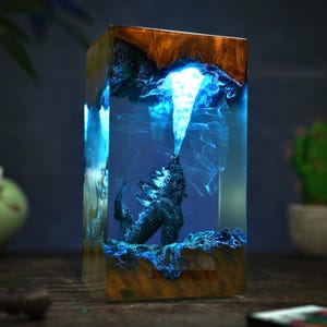 May include: A rectangular resin sculpture featuring a dark blue monster figure emitting a bright blue beam. The sculpture is encased in clear resin with a wooden base and top, creating a glowing effect. The overall theme is fantasy.