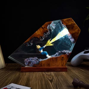 May include: A striking resin sculpture depicting a character in blue and white, unleashing a yellow energy attack. The artwork is set within clear resin, framed by wood, and mounted on a wooden base, creating a 3D effect.