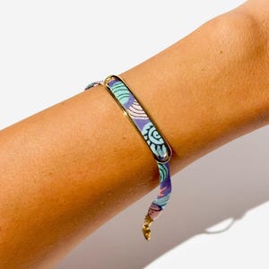 Könnte beinhalten: Ein goldfarbenes Metallarmband mit einem rechteckigen Mittelstück und einem gemusterten Stoffband. Der Stoff hat ein Design in Blau-, Grün- und Lilatönen. Das Armband wird mit einem goldfarbenen Verschluss befestigt. Das Armband ist ein Modeaccessoire.