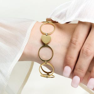May include: Gold-coloured bracelet with a heart-shaped charm and circular links. The bracelet has a delicate design with a small, clear accent on one of the links. The bracelet is shown on a wrist.