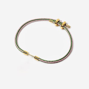May include: A circular bracelet featuring a braided cord of pink and green threads. The design includes polished gold-tone metal accents, specifically two sliding rings and a cylindrical connector piece that secures the ends of the cord together.