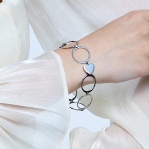 May include: Silver-toned bracelet featuring a heart-shaped charm and circular links. The bracelet is worn on a wrist, showcasing its design and metallic sheen. The jewelry is a stylish accessory.