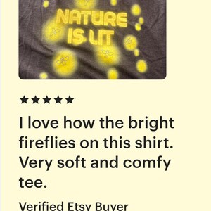 Op de afbeelding: Donkergrijs t-shirt met de gele tekst "NATURE IS LIT", omringd door gele vuurvlieggraphics. Het t-shirt heeft een ronde hals.