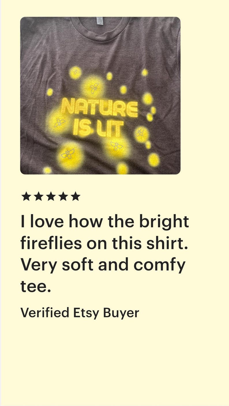Op de afbeelding: Donkergrijs t-shirt met de gele tekst "NATURE IS LIT", omringd door gele vuurvlieggraphics. Het t-shirt heeft een ronde hals.