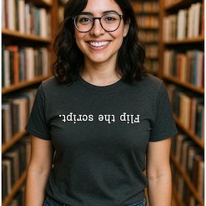 May include: A woman smiles, wearing a dark grey t-shirt with the words "Flip the script." She is in front of a bookshelf. She wears glasses and blue jeans.
