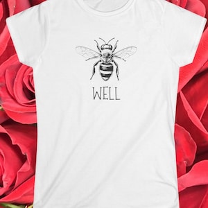 Op de afbeelding: Wit T-shirt met een zwart-witte illustratie van een bij. Onder de bij staat het woord "WELL" in een strak lettertype. De tee heeft een ronde hals en korte mouwen.