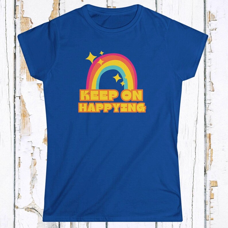 Op de afbeelding: Blauw T-shirt met een kleurrijk regenboogontwerp, gele sterren en de tekst "KEEP ON HAPPYING" in oranje letters. Het shirt heeft een ronde hals en korte mouwen.