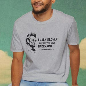 May include: A heather gray t-shirt featuring a black and white portrait of Abraham Lincoln. The shirt displays the quote, "I walk slowly but I never walk backward" attributed to Abraham Lincoln.