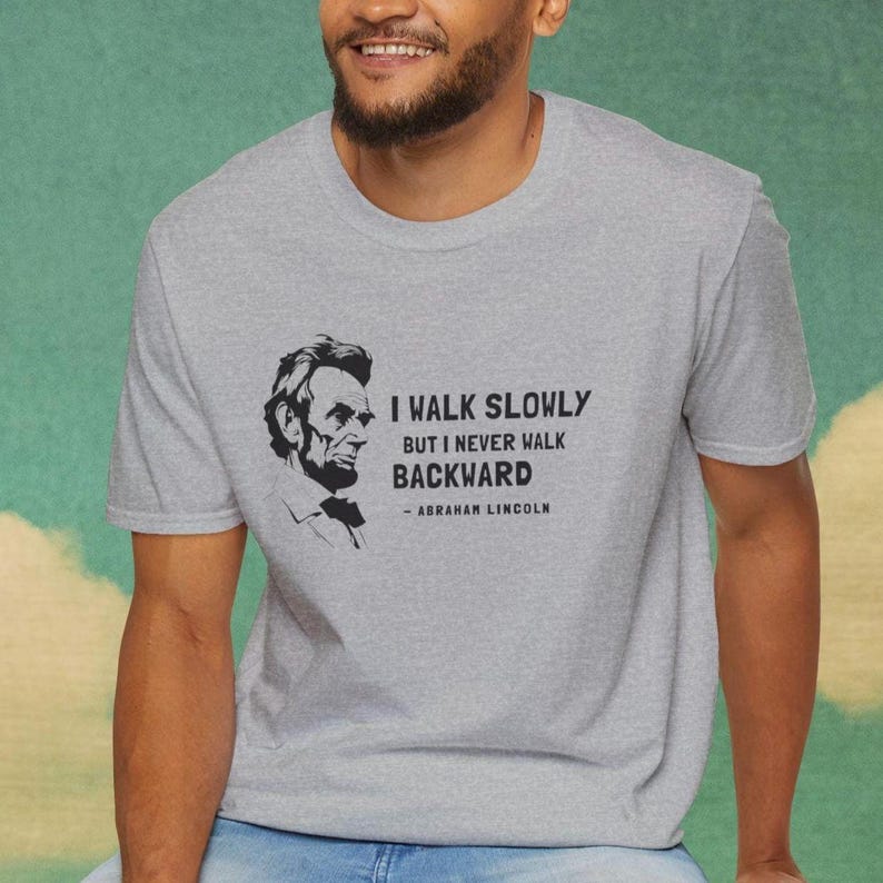 May include: A heather gray t-shirt featuring a black and white portrait of Abraham Lincoln. The shirt displays the quote, "I walk slowly but I never walk backward" attributed to Abraham Lincoln.