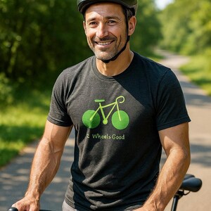 May include: A man wearing a black bicycle helmet and a dark gray t-shirt with a green bicycle graphic and the text "2 Wheels Good." He is holding a black bicycle on a paved path with trees in the background.