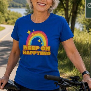 Op de afbeelding: Een koningsblauw t-shirt met een regenboog- en sterrenafbeelding, en de woorden "KEEP ON HAPPYING" in geel en oranje. Het shirt wordt gedragen door een persoon die naast een fiets staat.