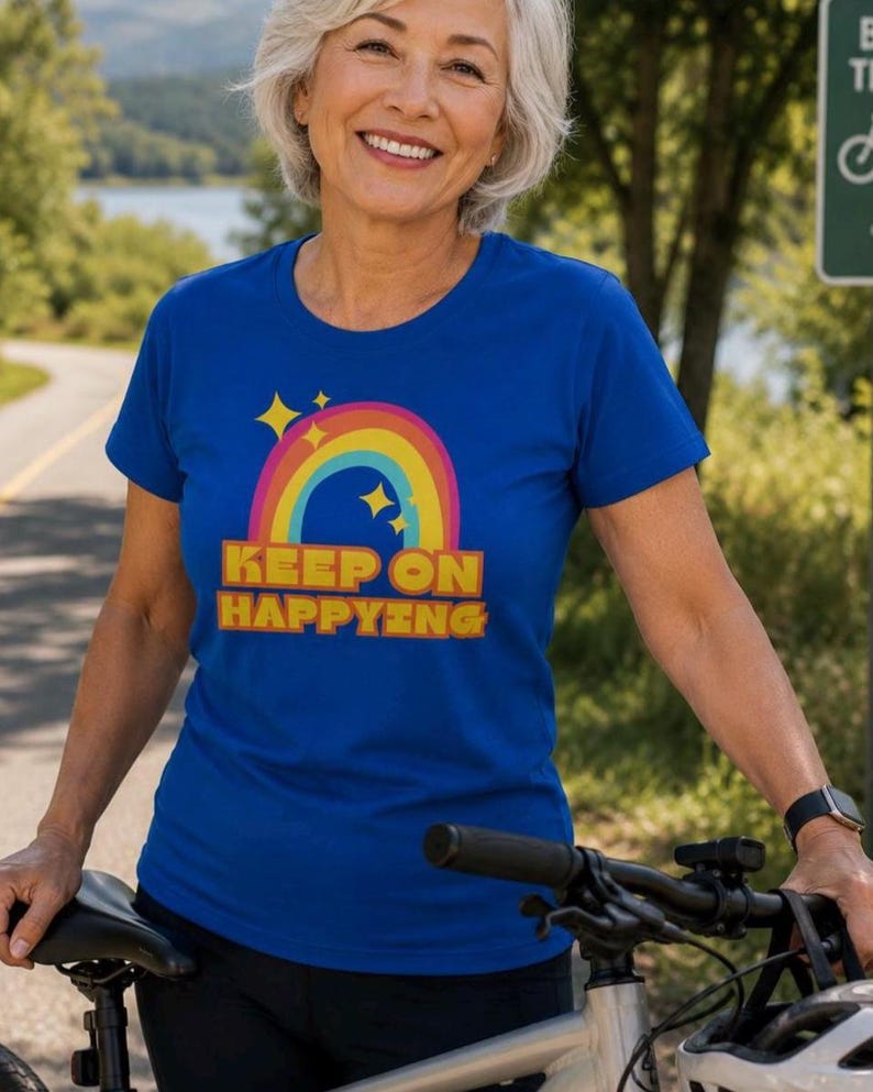Op de afbeelding: Een koningsblauw t-shirt met een regenboog- en sterrenafbeelding, en de woorden "KEEP ON HAPPYING" in geel en oranje. Het shirt wordt gedragen door een persoon die naast een fiets staat.