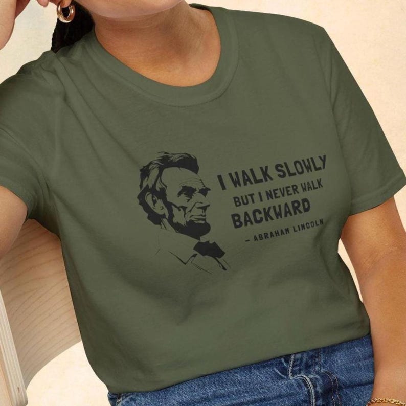 May include: Olive green t-shirt featuring a black graphic of Abraham Lincoln and the quote, "I walk slowly but I never walk backward." The quote is attributed to Abraham Lincoln. The shirt is a crew neck style.
