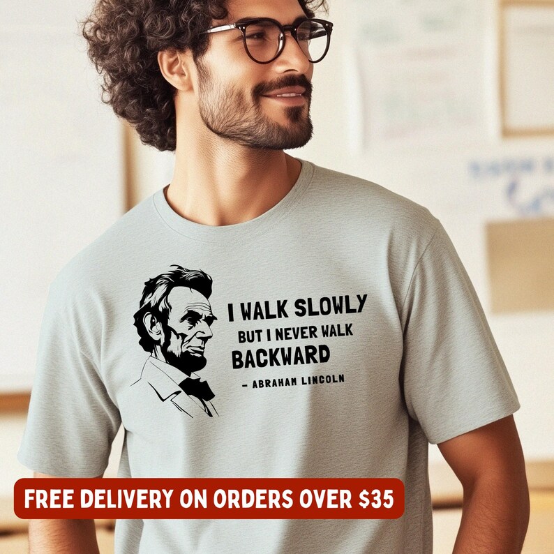 May include: A light gray t-shirt featuring a black and white portrait of Abraham Lincoln and the quote, "I walk slowly but I never walk backward." The text is in a bold, easy-to-read font. The shirt is a crew neck style.