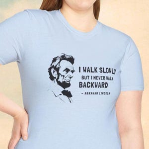 May include: Light blue t-shirt featuring a black and white portrait of Abraham Lincoln. The shirt displays the quote "I walk slowly but I never walk backward" attributed to Lincoln.