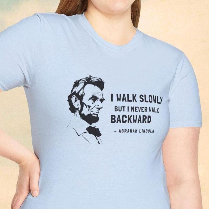 May include: Light blue t-shirt featuring a black and white portrait of Abraham Lincoln. The shirt displays the quote "I walk slowly but I never walk backward" attributed to Lincoln.