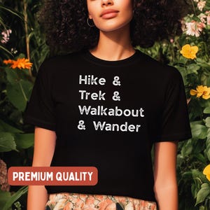 K&ouml;nnte beinhalten: Schwarzes T-Shirt mit den wei&szlig;en Worten &bdquo;Hike & Trek & Walkabout & Wander&ldquo;. Das Shirt hat auch ein &bdquo;Premium Quality&ldquo;-Etikett in einem roten Rechteck. Das Shirt wird vor einem Hintergrund aus gr&uuml;nem Laub und gelben Blumen getragen.