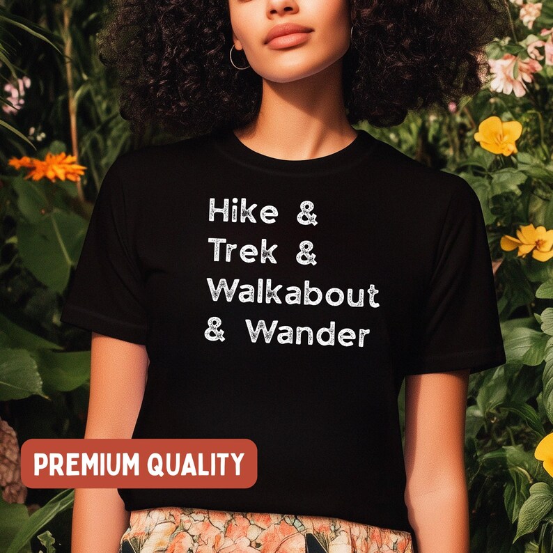 K&ouml;nnte beinhalten: Schwarzes T-Shirt mit den wei&szlig;en Worten &bdquo;Hike & Trek & Walkabout & Wander&ldquo;. Das Shirt hat auch ein &bdquo;Premium Quality&ldquo;-Etikett in einem roten Rechteck. Das Shirt wird vor einem Hintergrund aus gr&uuml;nem Laub und gelben Blumen getragen.