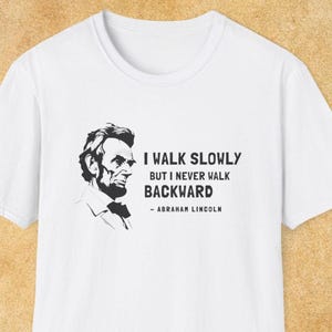 May include: White t-shirt with a black and white graphic of Abraham Lincoln's portrait. The text reads 'I WALK SLOWLY BUT I NEVER WALK BACKWARD - ABRAHAM LINCOLN'. The shirt has a crew neck and short sleeves. The background is a light tan color.