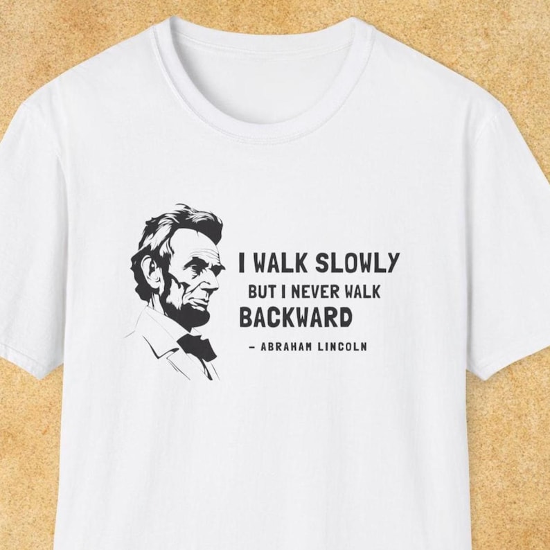 May include: White t-shirt with a black and white graphic of Abraham Lincoln's portrait. The text reads 'I WALK SLOWLY BUT I NEVER WALK BACKWARD - ABRAHAM LINCOLN'. The shirt has a crew neck and short sleeves. The background is a light tan color.