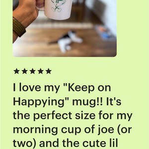 Op de afbeelding: Witte keramische mok met de tekst "KEEP ON HAPPYING" en een groen stripfiguur van koffie. De mok wordt door een persoon vastgehouden. Een leuke, motiverende koffiemok.