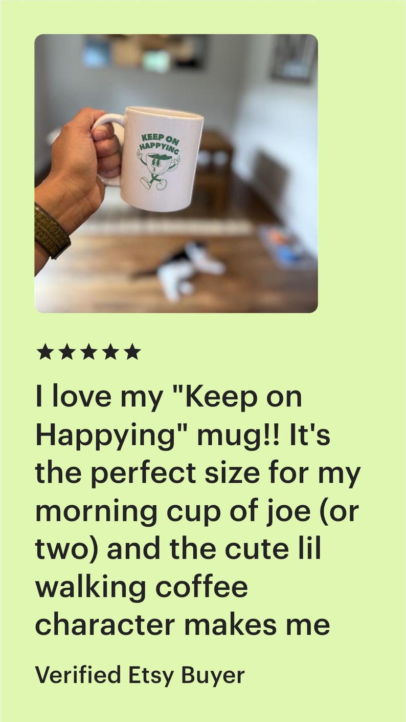 Op de afbeelding: Witte keramische mok met de tekst "KEEP ON HAPPYING" en een groen stripfiguur van koffie. De mok wordt door een persoon vastgehouden. Een leuke, motiverende koffiemok.