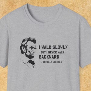 May include: A heather gray t-shirt featuring a black and white portrait of Abraham Lincoln. The shirt includes the quote, "I WALK SLOWLY BUT I NEVER WALK BACKWARD" attributed to Abraham Lincoln.