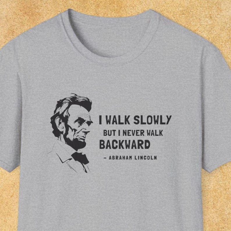 May include: A heather gray t-shirt featuring a black and white portrait of Abraham Lincoln. The shirt includes the quote, "I WALK SLOWLY BUT I NEVER WALK BACKWARD" attributed to Abraham Lincoln.