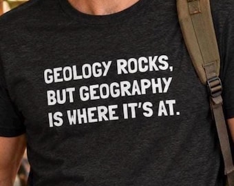 May include: A dark charcoal grey short-sleeved t-shirt featuring white text printed on the chest that reads: GEOLOGY ROCKS, BUT GEOGRAPHY IS WHERE IT'S AT. The shirt has a crew neckline and is worn with a brown backpack strap visible over the shoulder.