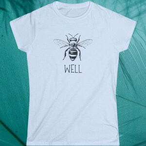 Op de afbeelding: Wit t-shirt met een zwart-witte illustratie van een gedetailleerde bij. Onder de bij staat het woord "WELL". Het t-shirt heeft een ronde hals en korte mouwen. De afbeelding toont het ontwerp van de bij.