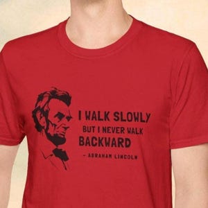 May include: Red t-shirt featuring a black silhouette of Abraham Lincoln and the quote, "I walk slowly but I never walk backward." The quote is attributed to Abraham Lincoln. The shirt is a crew neck.