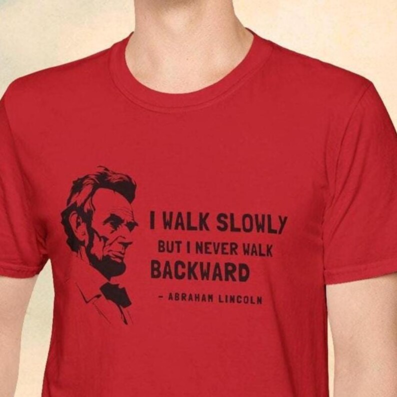 May include: Red t-shirt featuring a black silhouette of Abraham Lincoln and the quote, "I walk slowly but I never walk backward." The quote is attributed to Abraham Lincoln. The shirt is a crew neck.