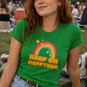Op de afbeelding: Een groen t-shirt met een regenboog- en sterrenontwerp en de tekst "KEEP ON HAPPYING" in geel en oranje. Het shirt wordt gedragen met een spijkerbroekshort en een bruine riem. De persoon glimlacht.