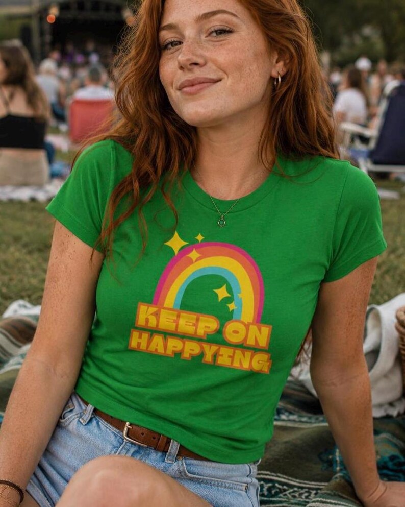 Op de afbeelding: Een groen t-shirt met een regenboog- en sterrenontwerp en de tekst "KEEP ON HAPPYING" in geel en oranje. Het shirt wordt gedragen met een spijkerbroekshort en een bruine riem. De persoon glimlacht.