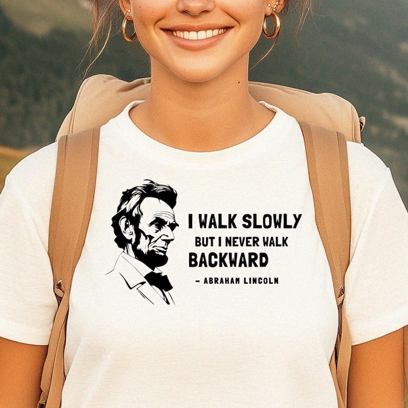 May include: White t-shirt featuring a black and white portrait of Abraham Lincoln with the quote "I walk slowly but I never walk backward." The shirt is a classic crew neck style.