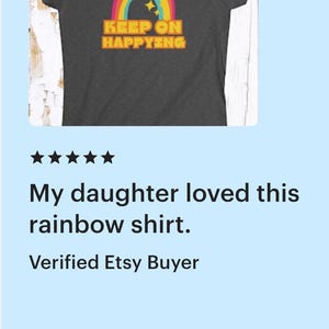 Op de afbeelding: Donkergrijs t-shirt met een kleurrijke regenboogafbeelding en de tekst "KEEP ON HAPPYING". Het regenboogontwerp bevat rode, oranje, gele, groene en blauwe bogen, met gele sterren erboven. Een casual t-shirt.