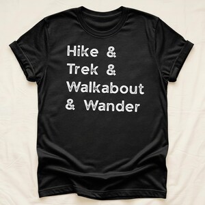 K&ouml;nnte beinhalten: Schwarzes T-Shirt mit wei&szlig;em Text: "Hike & Trek & Walkabout & Wander." Das Shirt hat hochgekrempelte &Auml;rmel und wird vor einem neutralen Hintergrund pr&auml;sentiert. Der Text ist in einem l&auml;ssigen, handschriftlichen Stil gehalten.
