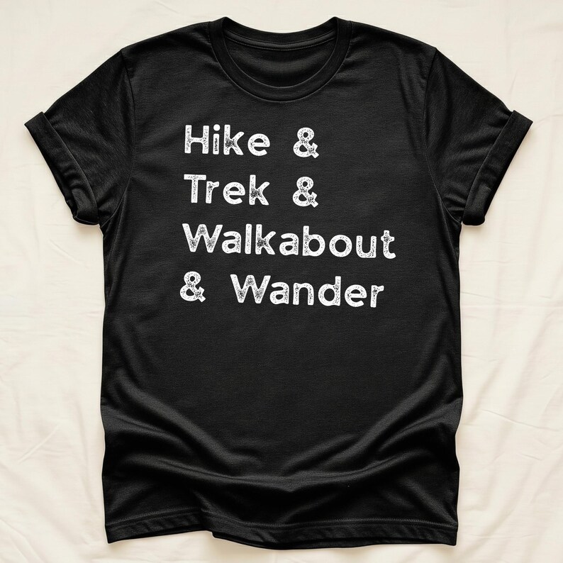K&ouml;nnte beinhalten: Schwarzes T-Shirt mit wei&szlig;em Text: "Hike & Trek & Walkabout & Wander." Das Shirt hat hochgekrempelte &Auml;rmel und wird vor einem neutralen Hintergrund pr&auml;sentiert. Der Text ist in einem l&auml;ssigen, handschriftlichen Stil gehalten.