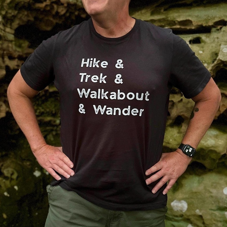 K&ouml;nnte beinhalten: Schwarzes T-Shirt mit dem wei&szlig;en Aufdruck "Hike & Trek & Walkabout & Wander". Das kurz&auml;rmelige T-Shirt scheint aus einem weichen Material zu sein. Die Person, die das T-Shirt tr&auml;gt, hat die H&auml;nde in die H&uuml;ften gestemmt.