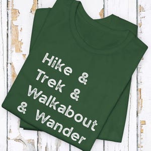 K&ouml;nnte beinhalten: Dunkelgr&uuml;nes T-Shirt mit wei&szlig;em Text: "Hike & Trek & Walkabout & Wander". Das gefaltete Shirt liegt auf einer wei&szlig;en Holzoberfl&auml;che. Der Text ist in einer serifenlosen Schriftart. F&uuml;r Wanderfreunde.