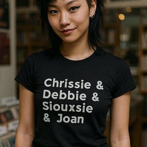 Op de afbeelding: Zwart T-shirt met witte tekst: "Chrissie & Debbie & Siouxsie & Joan". Het shirt met korte mouwen lijkt van een zacht materiaal te zijn. De persoon die het shirt draagt, glimlacht.