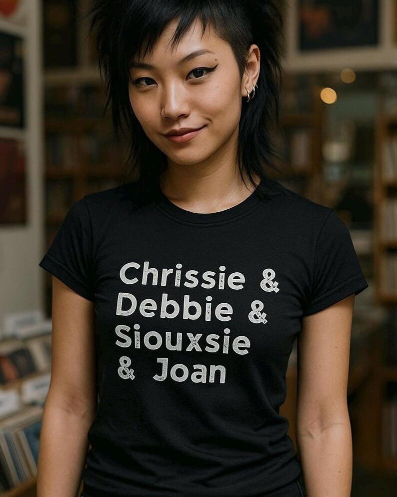 Op de afbeelding: Zwart T-shirt met witte tekst: "Chrissie & Debbie & Siouxsie & Joan". Het shirt met korte mouwen lijkt van een zacht materiaal te zijn. De persoon die het shirt draagt, glimlacht.
