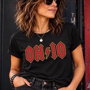 May include: A black short-sleeved t-shirt featuring a graphic print on the chest with the text OH-IO in a bold red font with a yellow outline. A yellow lightning bolt symbol separates the letters OH and IO in the center of the design.