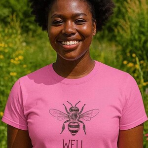 Op de afbeelding: Een roze T-shirt met een gedetailleerde zwart-witte illustratie van een bij en het woord "WELL" eronder. Het T-shirt heeft een ronde hals.
