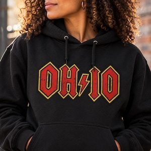 May include: A black hooded sweatshirt featuring a graphic print on the chest. The design displays the text OH-IO in a distressed red font with a yellow outline, incorporating a yellow lightning bolt symbol between the letters H and I.