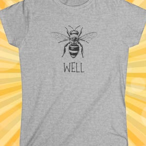 Op de afbeelding: Een lichtgrijs t-shirt met een zwart-witte illustratie van een bij met uitgespreide vleugels. Het woord "WELL" staat eronder in een eenvoudig lettertype. Het shirt heeft een ronde hals en korte mouwen.