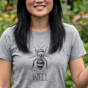 Op de afbeelding: Grijs t-shirt met een zwart-wit illustratie van een bij en het woord "WELL" eronder. Het t-shirt heeft een ronde hals.