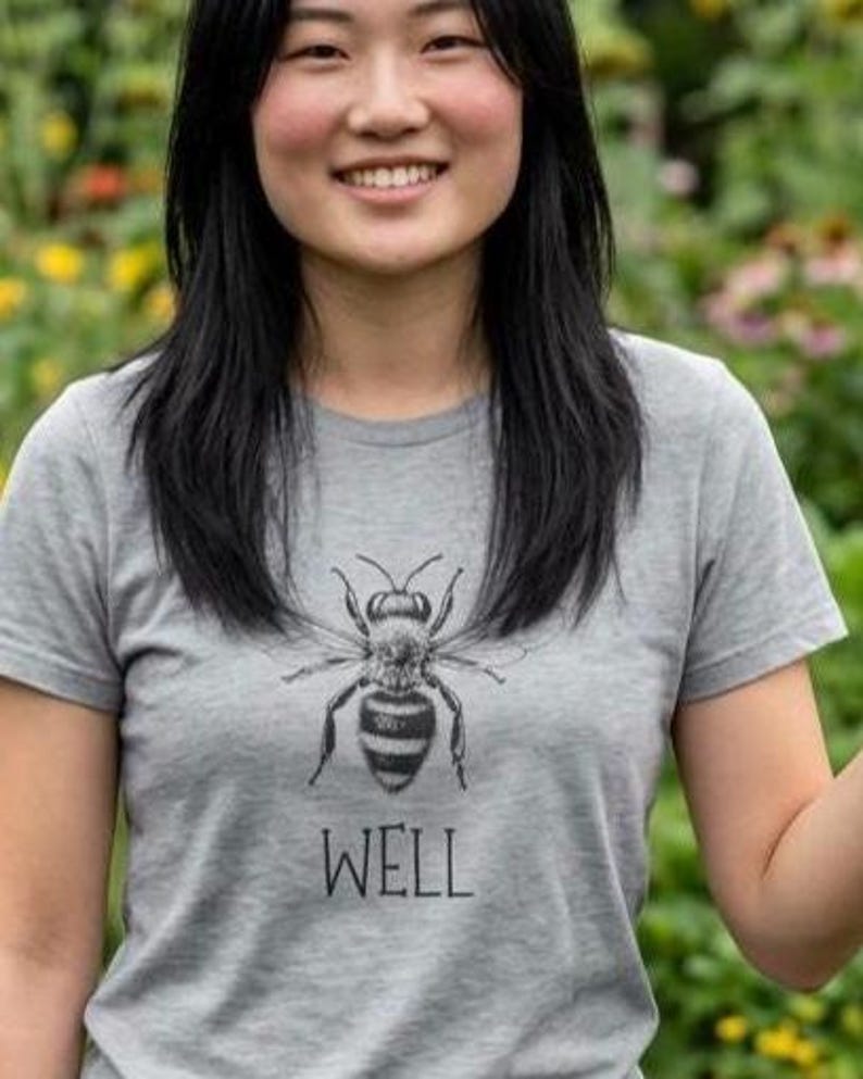 Op de afbeelding: Grijs t-shirt met een zwart-wit illustratie van een bij en het woord "WELL" eronder. Het t-shirt heeft een ronde hals.